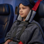 A child wearing a dark blue sleep mask and a gray fleece blanket sits in an airplane seat with a red and gray neck cradle and black seatbelt harness.