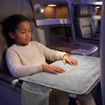 A child sits in a premium airplane cabin, resting their head on a light gray fleece headrest cover attached to a fold-out tray table. The child is wearing a cream knit sweater, and the cabin interior features blue ambient lighting.